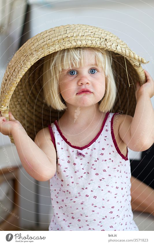 Blonde, smart, roll-eyed girl in a flowered undershirt, puts a large, beige basket made of natural materials over her head and looks into the camera. Funny, funny, imaginative, creative children's game at home, in the bright apartment.