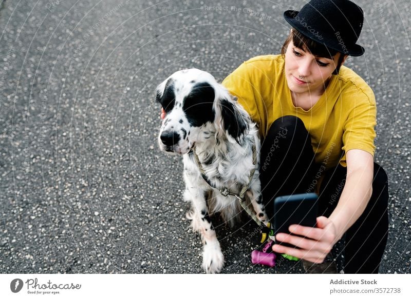 Young lady using smartphone while sitting on ground with dog woman walk selfie fun park nature hipster subculture style together companion street hat memory