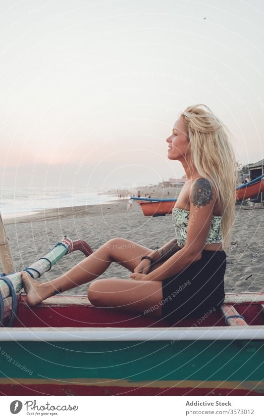 Cheerful lady resting on wooden boat on sandy beach woman ocean coast sunset shore calm harmony solitude peace water bench positive young casual dress summer