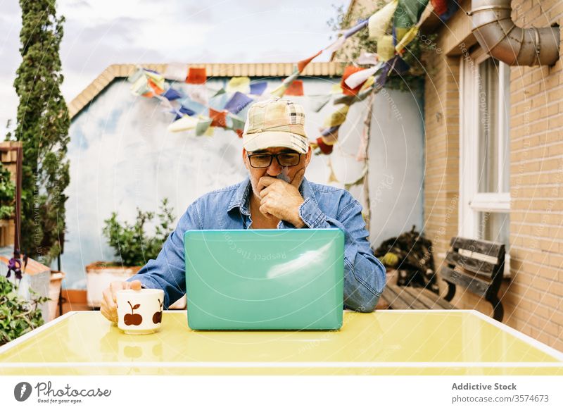 Pensive senior man in glasses having hot drink and using laptop on terrace of country house pensive read watch mug thoughtful device coffee internet male