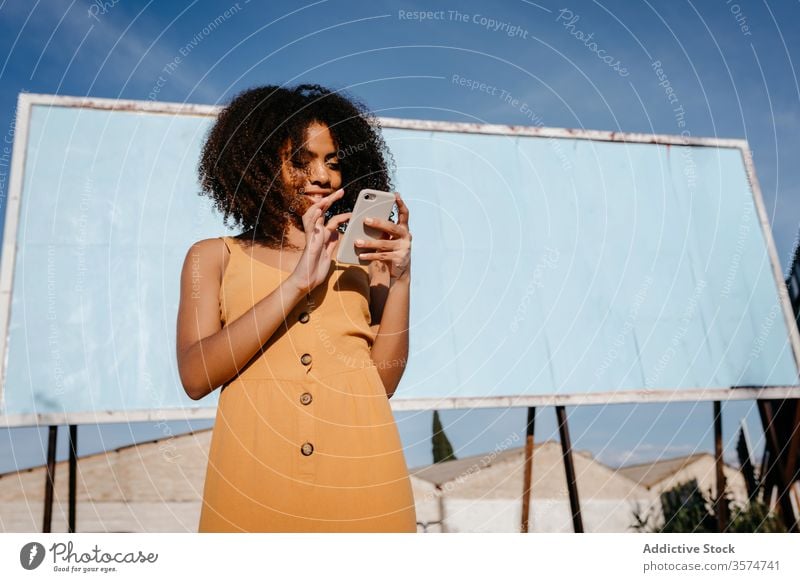 Young satisfied ethnic woman using cellphone on street against empty big board in suburb smartphone billboard use joy browsing surfing read banner social media