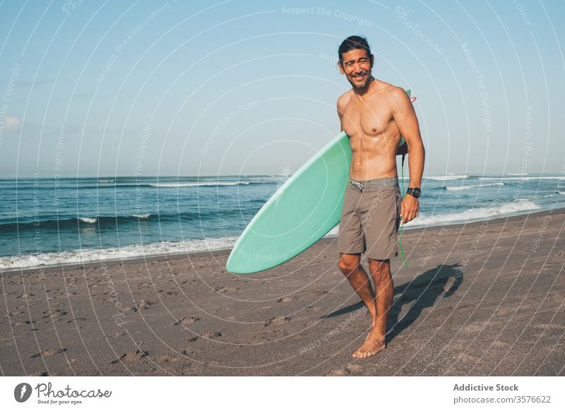 Cheerful surfer with surfboard standing on beach man sea happy active ocean coast smile shirtless ethnic male lifestyle sand summer shore vacation water