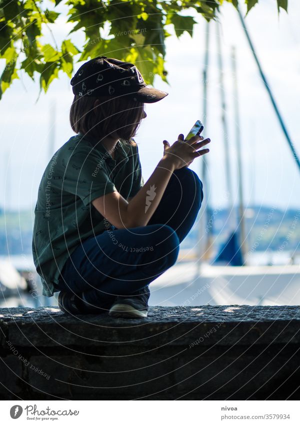 Junge spielt auf dem Handy am Hafen junge handy smartphone sitzen sommer game spielen Cap Schatten blau Natur Sommer Pflanze See Landschaft Colour photo Himmel