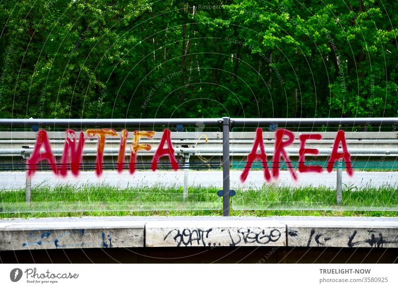 Literally: | Good news...  Graffiti ANTIFA AREA in red capital letters on a transparent partition wall of a tram stop at a highway with forest in the background