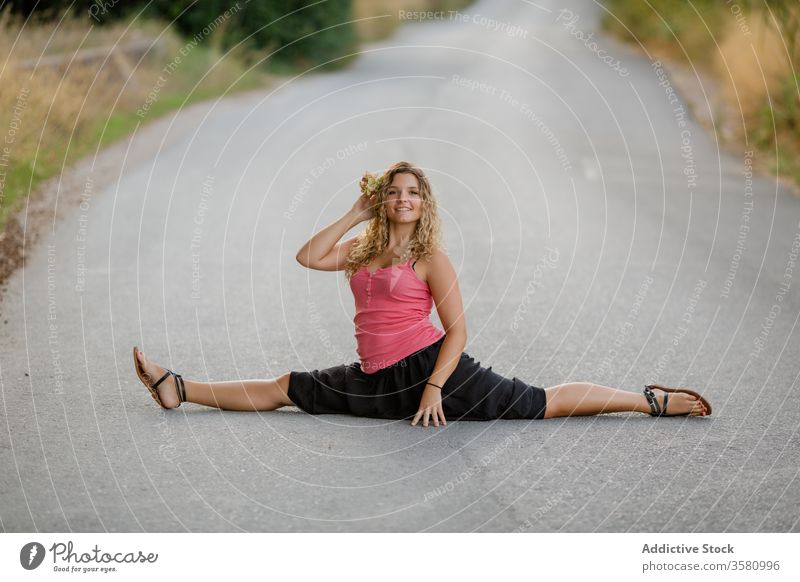 Cheerful woman doing splits on road in countryside stretch flexible cheerful smile healthy asphalt female pose practice balance casual flower hair wellness