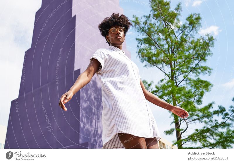 Calm black woman on street against colorful violet exterior of skyscraper and green tree in city dress appearance personality facade calm pensive style glasses