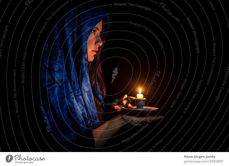 Sad young woman with candle and blue headscarf Face sad black portrait frontal profile serious art gloomy candlestick hands flame burn light scary look eyes