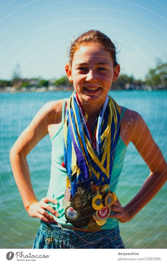 My champion. // A girl is standing happily at the edge of the river, laughing. She has many medals hanging around her neck. She has waved her arms. Girl