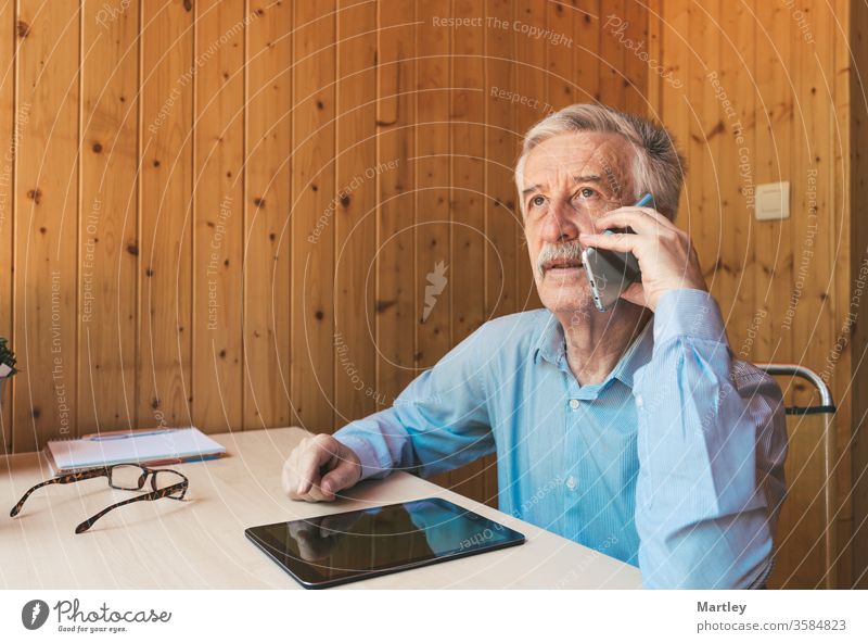Grandfather talking to his grandchildren and relatives, who were quarantined from home, about the coronavirus. Elderly man using smartphone and tablet in his office