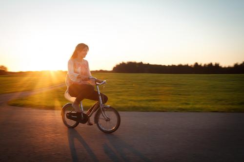 Long-haired, brunette teenagers, riding a small bicycle on the street in the evening light. Cycling in the twilight, passing green meadows and woods, the teenager, like a funny giant on the oversized, tiny dwarf bike.