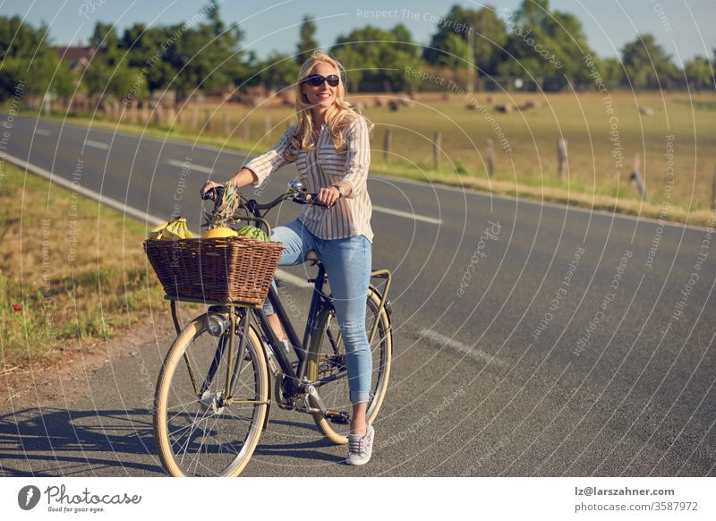 Middle-aged blond woman shopping for groceries on her bicycle stopped at the side of the road with a basketful of healthy fresh produce smiling as she faces into the spring sunshine in a rural landscape