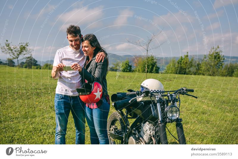 Couple looking mobile with a motorcycle happy couple smartphone picture video trip man smiling cell phone embracing motorcycle helmet custom attractive built