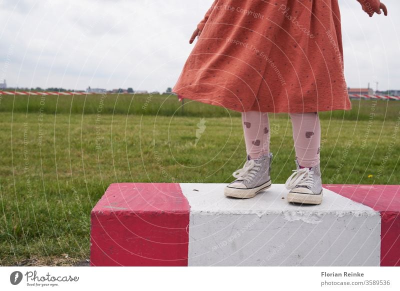 Girl with dress and sneakers stands on a red and white striped concrete barrier on the Tempelhofer Feld, in the background a meadow and red and white barrier tape