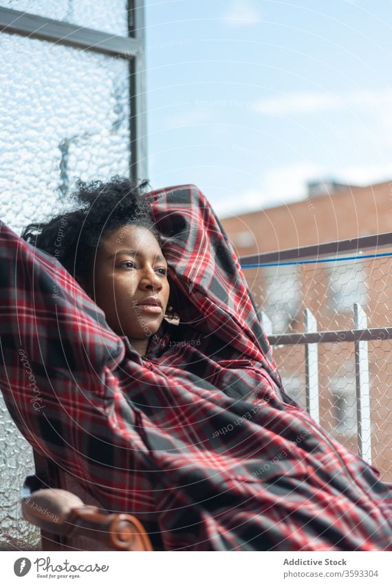 Dreamy black woman relaxing in armchair on balcony thoughtful dreamy checkered shirt pensive rest think female ethnic african american sit comfort cozy tranquil