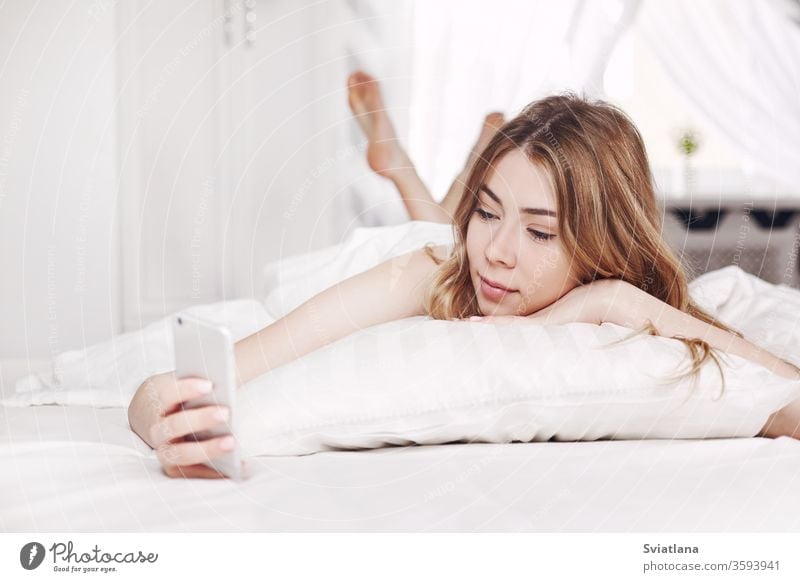 Cute smiling girl looks at the phone, lying on the bed. A girl reads messages on her phone when she wakes up in the morning. woman asleep home pillow bedtime