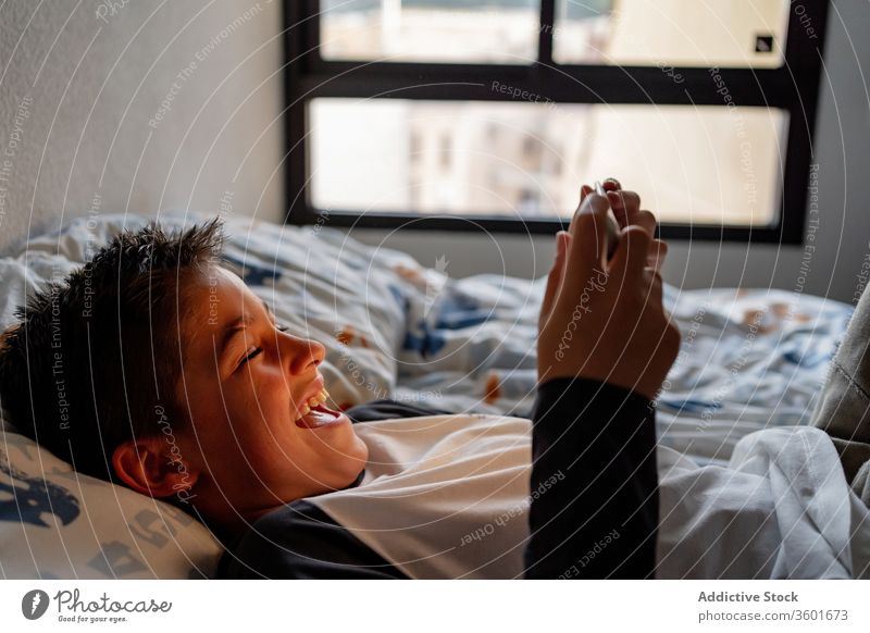 Delighted boy using tablet in bedroom during weekend kid blanket night watching cheerful pajama home adorable device gadget child internet browsing cozy rest
