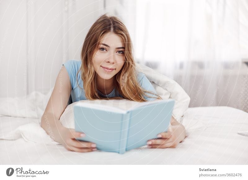 Beautiful girl in blue pajamas reading a book, lying on the bed in the bedroom. Training, education, Hobbies. reads home leisure young woman beautiful resting