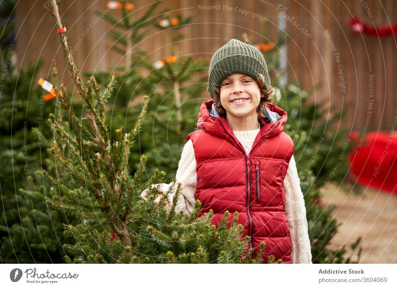Beautiful blond boy with green wool hat, red vest, white pullover, blue pants and yellow boots choosing his Christmas tree fresh closeup boys december holiday