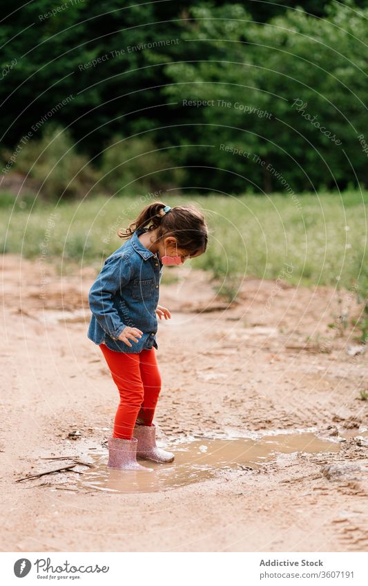 Curious girl surgical mask playing in mud puddle child having fun curious kid rubber boot cute dirty water wet adorable childhood joy activity playful ripple