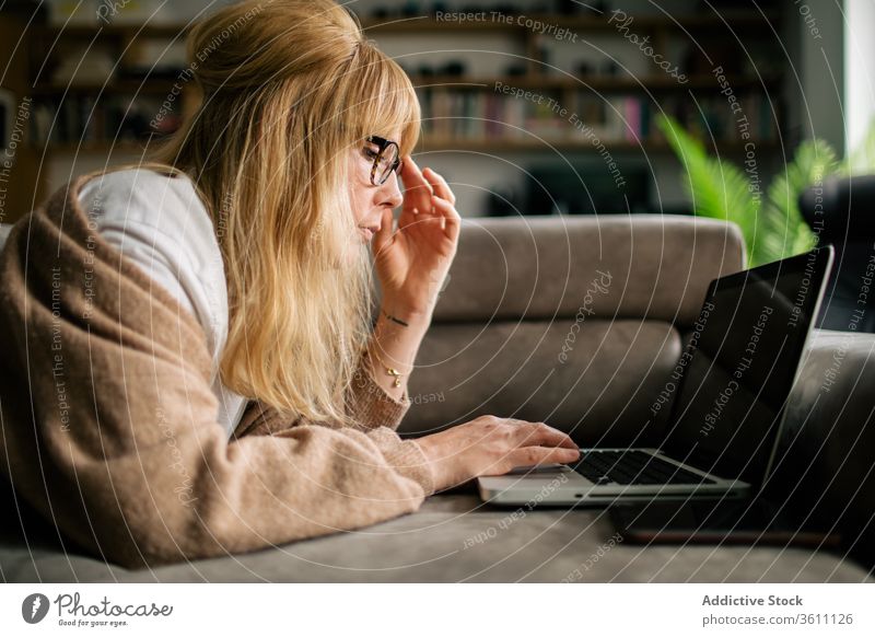 Pensive woman working on laptop at home browsing freelance project using telework lying sofa remote female serious eyeglasses gadget internet device computer