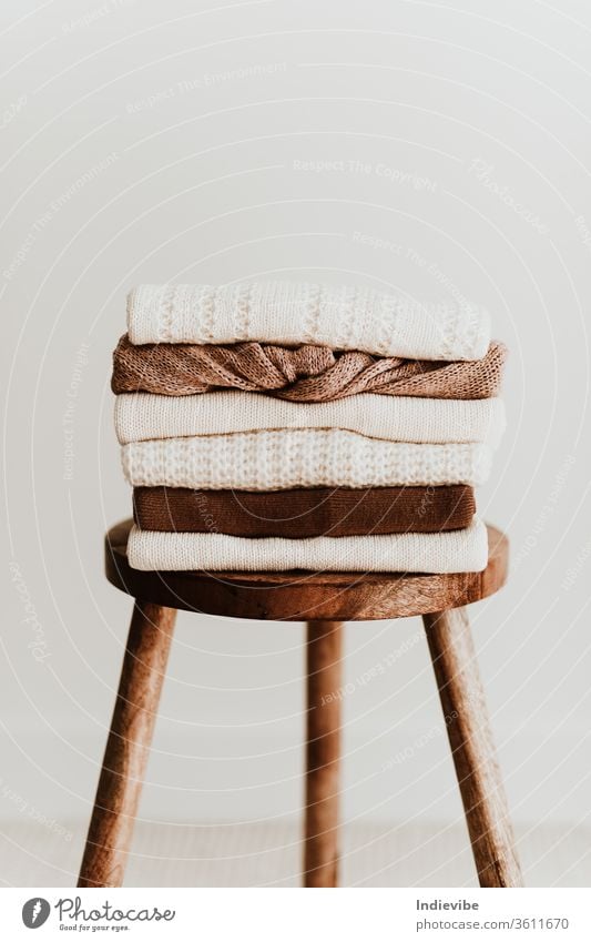 Women fashion composition. Warm winter female woollen knitted sweaters, pullovers stack on a wooden stool at white background. Modern clothes concept for magazine, blog, social media.