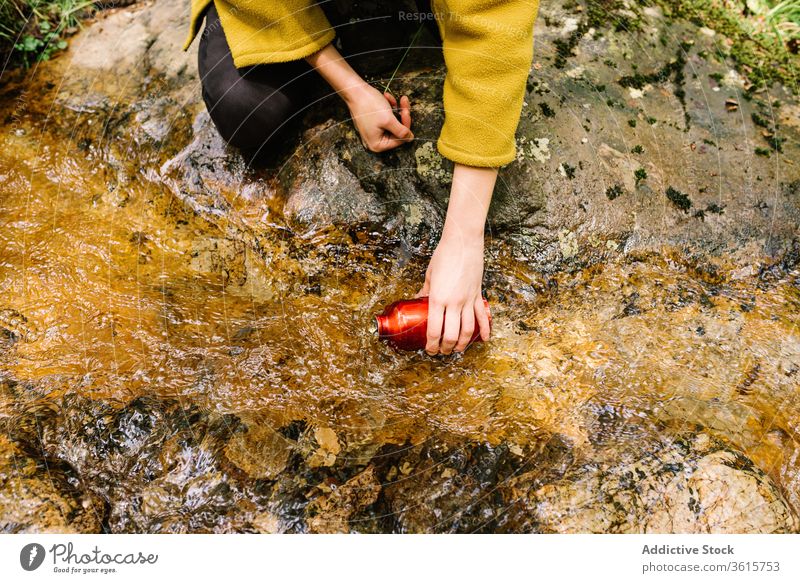 Anonymous woman taking water from brook in forest bottle clean style green female outerwear creek nature relax lifestyle countryside informal thirsty