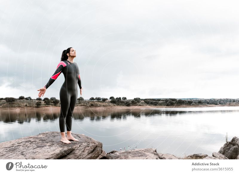 Inspired sportswoman on high rock against lake wetsuit diver water boulder evening inspiration sunset swimsuit nature shore hands on waist environment swimmer