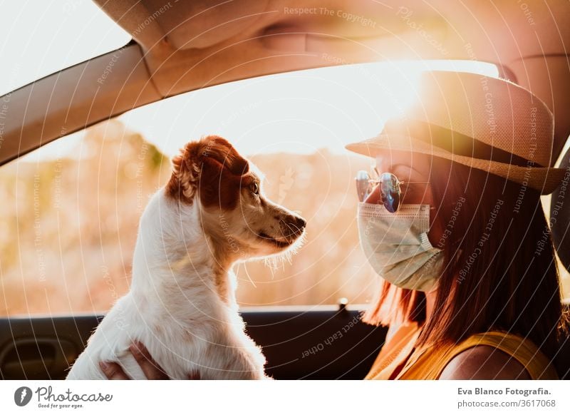 young woman in a car wearing protective mask, cuddling her cute small dog. Summer season. prevention corona virus concept sunset flare jack russell travel trip