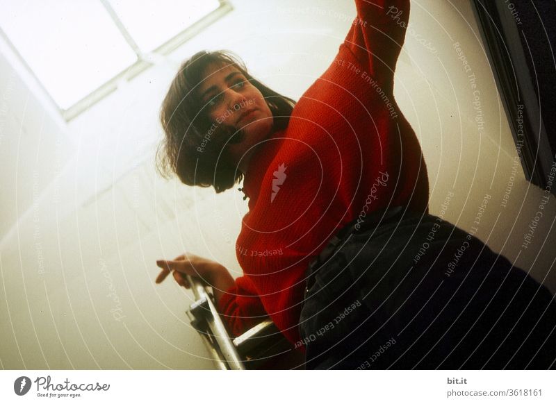 Young, pretty woman in red sweater stands on ladder in the attic under the bright window with light and makes selfie. Attractive model post in bright attic flat under skylight. Danger of falling from the household ladder in the household at home.