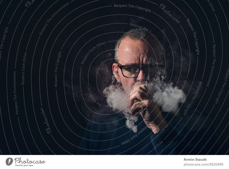 studio portrait of a man with vaporiser and glasses old beard gray vaping smoke cool serious e-cigarette electronic vapes male smoking blowing healthy equipment