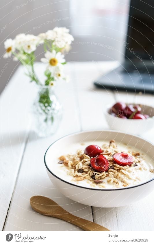 Muesli with yoghurt and cherries on a white wooden table and a laptop. Healthy breakfast. Cereal Breakfast Oat flakes Yoghurt Bowl Wooden door home office Spoon