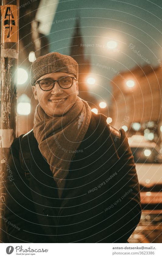 Young man in glasses grey cap black jacket and dark scarf on highway avenue looking in camera holding slightly smiling twilight evening night summer sight young