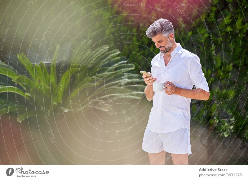 Brutal man browsing smartphone in backyard summer courtyard talk morning coffee handsome fresh male cellphone outfit gadget device drink beverage mobile call