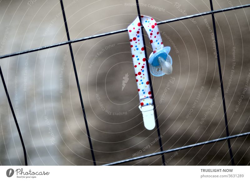 A sky-blue baby pacifier on a colourful ribbon hangs lonely and abandoned in a wire mesh in front of a dark blurred background Soother pacifier band Baby