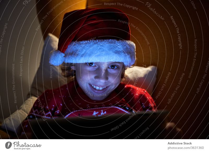 Happy kid enjoying new tablet on Christmas eve at home boy christmas santa hat holiday toothy smile internet online using gadget device reflection child
