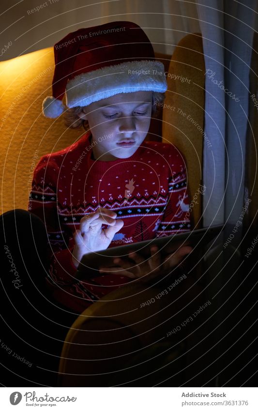 Kid enjoying new tablet on Christmas eve at home boy christmas santa hat holiday internet online using gadget device reflection child new year relax evening