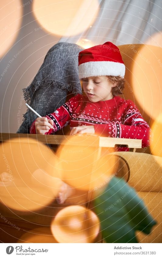 Boy in Christmas wear sitting in cozy armchair boy santa hat christmas holiday pencil happy comfort domestic sweater glad cheerful comfortable bokeh positive