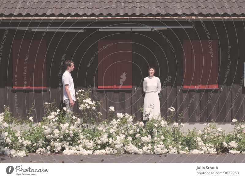 Bridal couple standing in front of a wooden hut bridal couple Man Woman White Retro Rose plants roses green Concrete Wood panelling shutters Closed Tiled roof