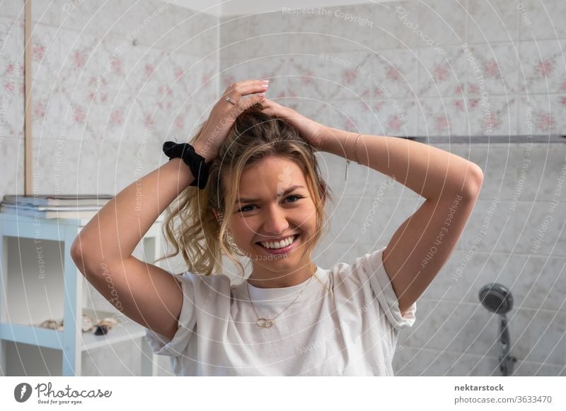 Young Woman Tying Back Hair and Smiling in Bathroom woman long hair hairstyle portrait smile lifestyle looking at camera blond hair young woman one woman only