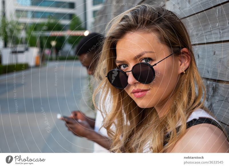 Young Woman with Round Sunglasses Looking at Camera woman portrait sunglasses blurred background smartphone lifestyle looking at camera 2 people male female
