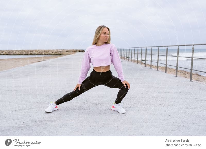 Fit sportswoman doing side lunge on embankment near ocean stretch leg workout activewear fence flexible training warm up sneakers wellbeing sky cloudy activity