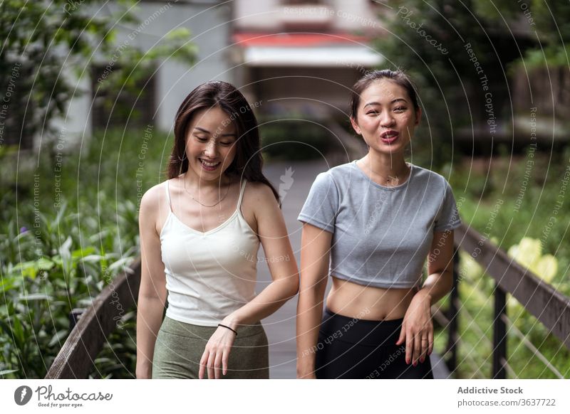 Positive young ethnic women walking along narrow bridge in countryside girlfriend positive chat toothy smile casual fit joy stroll communicate together