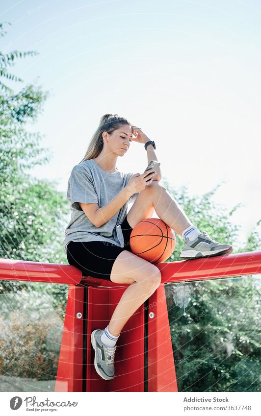 Sportswoman with basketball ball using smartphone on playground court serious young rest browsing mobile sport sportswoman athlete training activity player