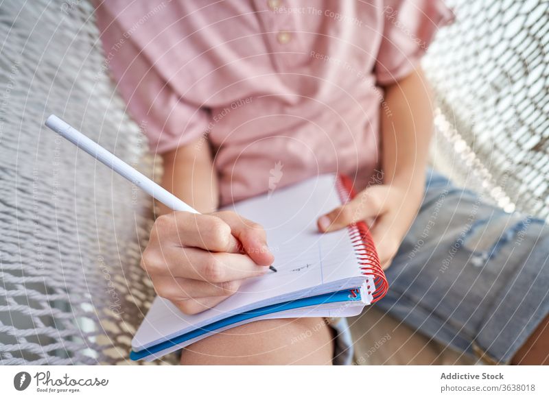 Anonymous schoolboy in eyeglasses studying in hammock with copybook at home education homework knowledge intelligent focused pencil childhood comfort harmony