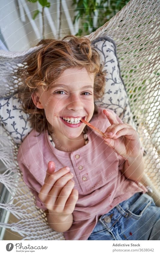 Content boy enjoying delicious fruit ice lolly sitting in hammock eat kid weekend legs crossed barefoot mouth opened idyllic childhood comfort rest harmony