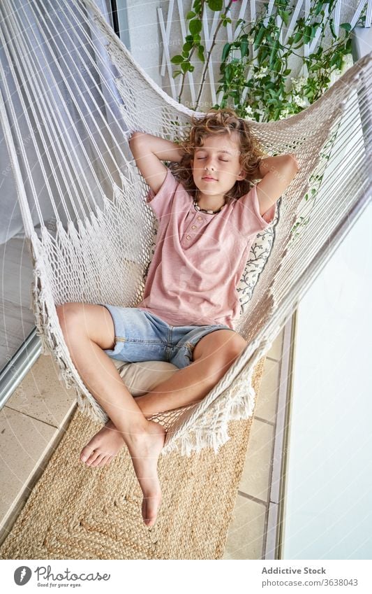 Peaceful boy sleeping in hammock on balcony at home eyes closed legs crossed lean on hand harmony idyllic rest barefoot child peace calm asleep weekend tranquil