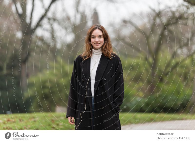 Young Woman with Brown Hair and Black Coat Smiling in Park caucasian ethnicity woman female earphones audio music listening brown hair real life model