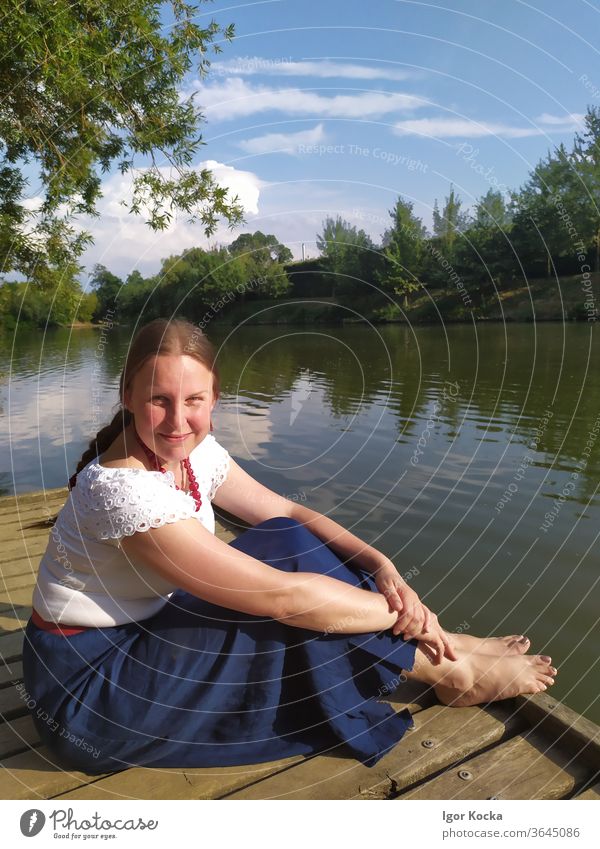 Portrait Of Smiling Woman Sitting On Pier Over Lake Pond pier sitting Sunlight side view portrait Looking into the camera adult Mature Trees Sky Nature