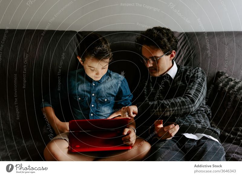 Schoolboy with brother browsing tablet on comfy sofa siblings using gadget contemporary relax rest chat legs crossed surfing connection cozy together point