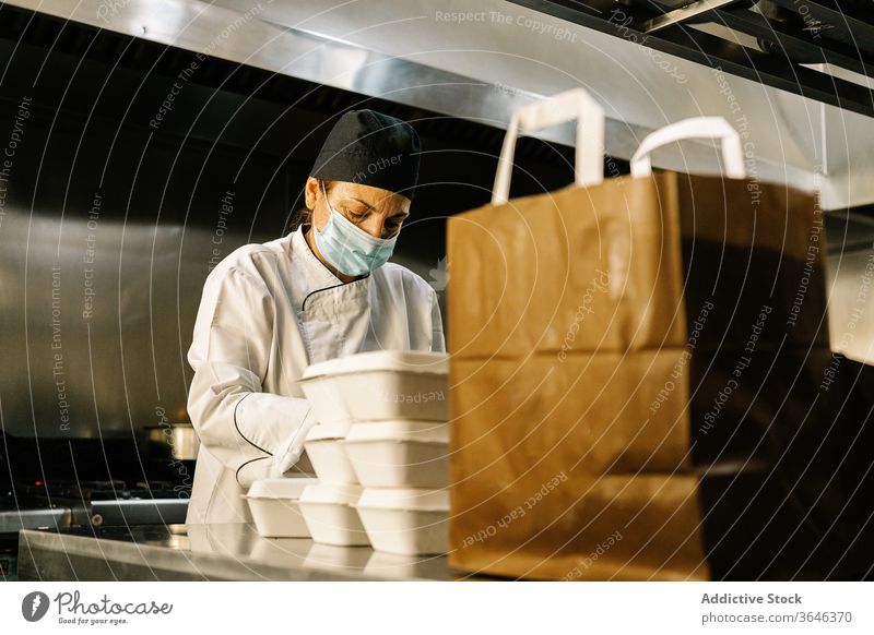 Focused female chef in respirator packing food for takeaway woman kitchen concentrate paper bag restaurant coronavirus focus uniform container occupation